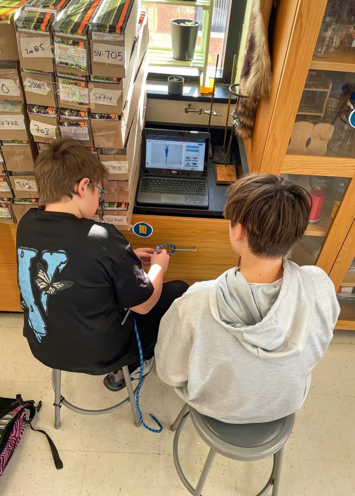 Two male middle school students sitting on stools, working together at a laptop on a table surrounded by storage boxes.