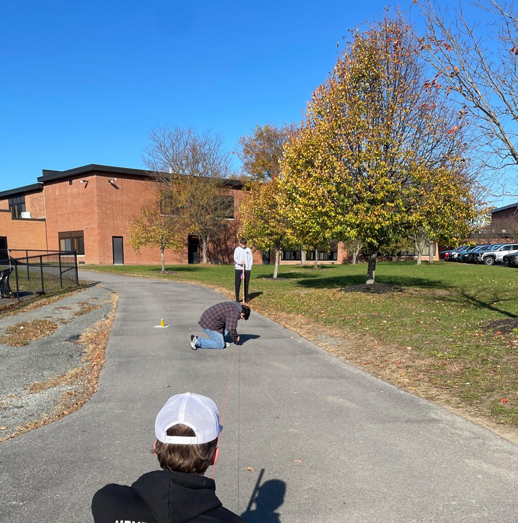 A student is kneeling in the middle of a gravel pathway measuring, while two people hold opposite ends of the tape measure. The school building is the background as well as some trees and a clear blue sky.