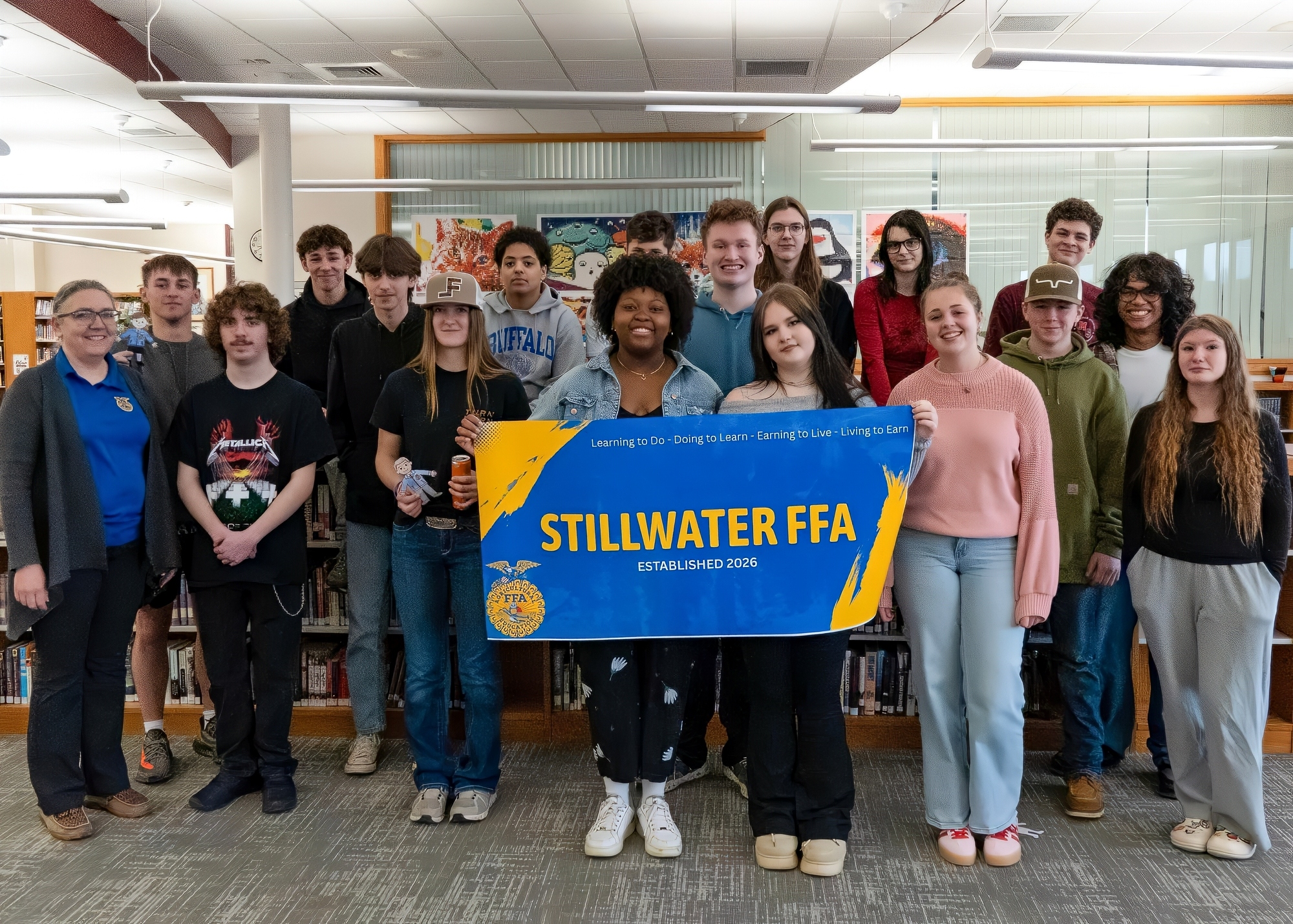 A group of students and their teacher gathered in the school library, holding a banner that reads "STILLWATER FFA". All are smiling for a photo.