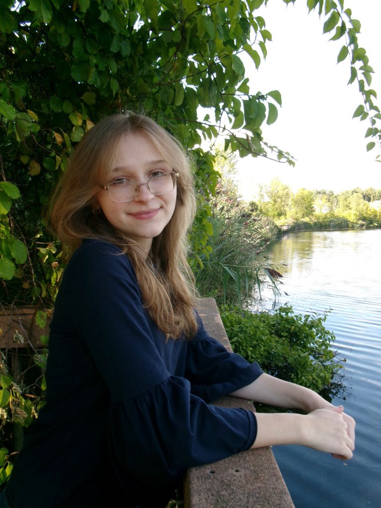 A young woman with long, light brown hair and glasses is leaning on a wooden railing beside a body of water, smiling at the camera. She is wearing a blue top, and there are trees and greenery in the background.