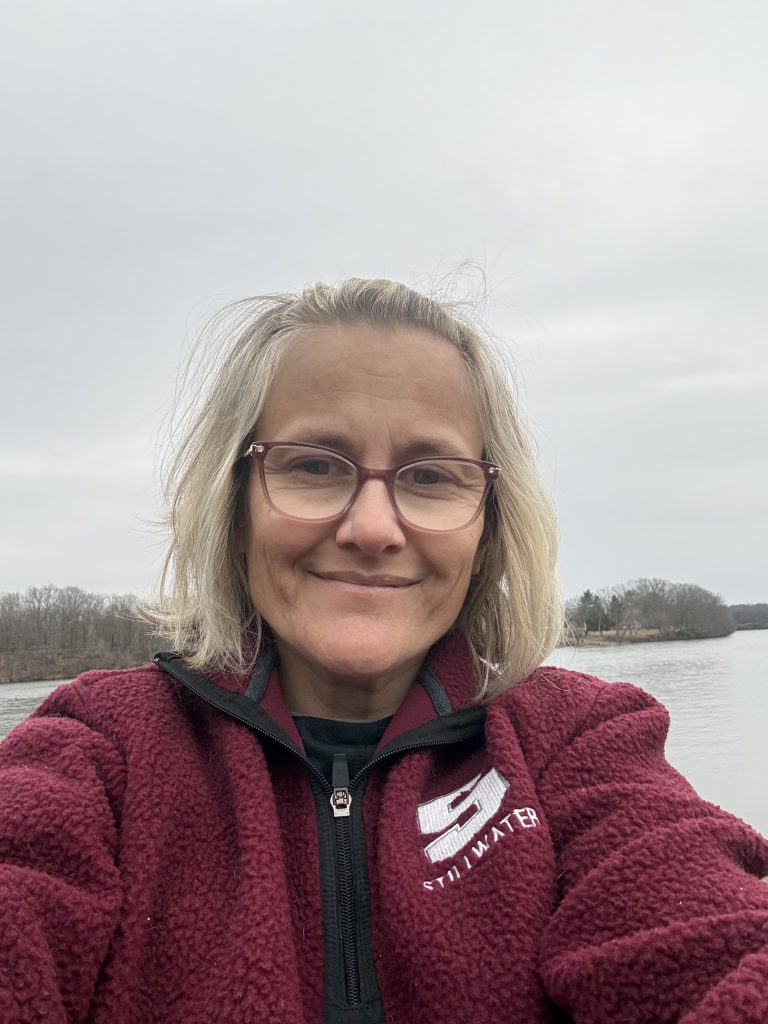 Woman with light blonde hair wearing glasses and a maroon sweatshirt with the white Stillwater "S" logo, smiling.