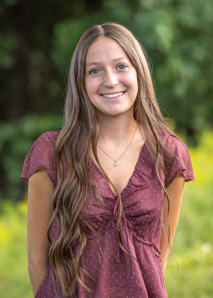 A young woman with long brown hair in a maroon top smiling stands outdoors against a lush, green background.