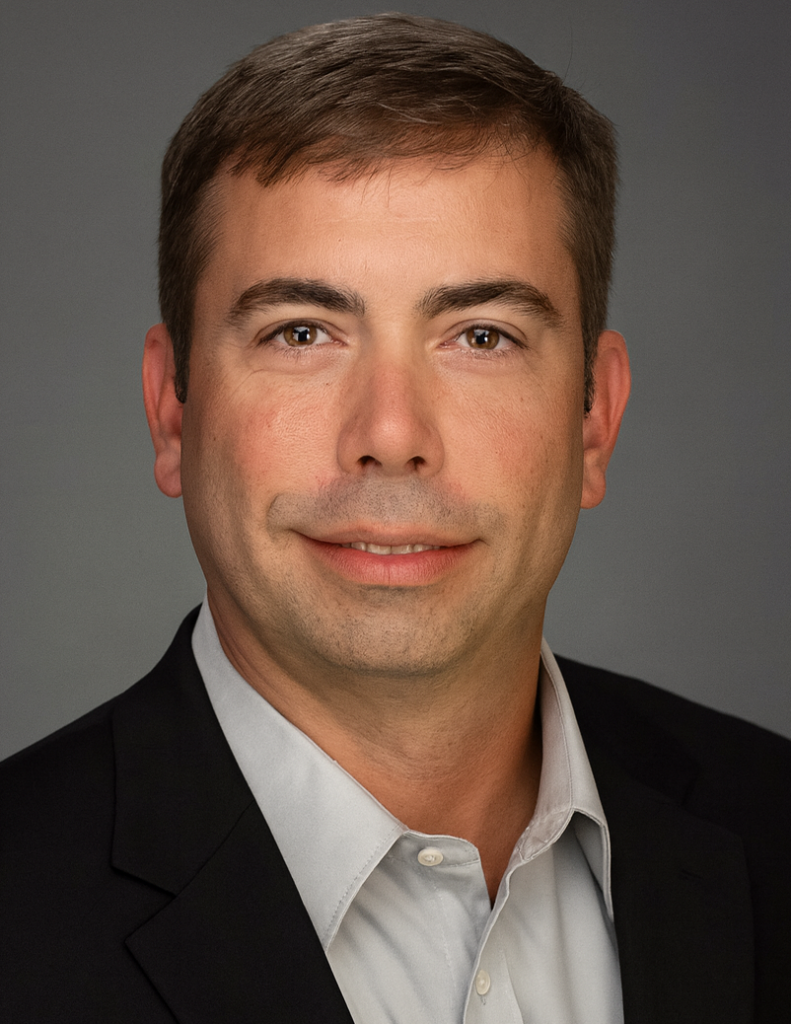 Man with brown short hair wearing a black coat and gray collared shirt, smiling towards the camera.