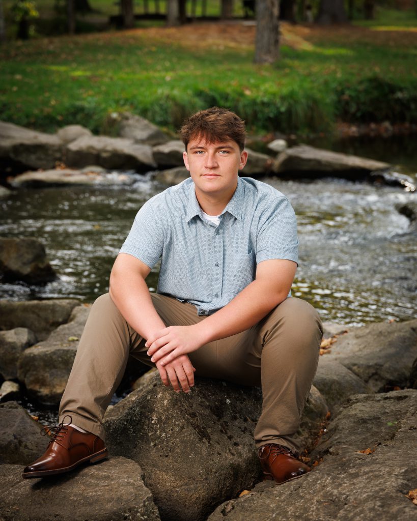 A young man in a light blue polo shirt and tan pants sits on a large rock by a stream. The background features lush greenery and trees.