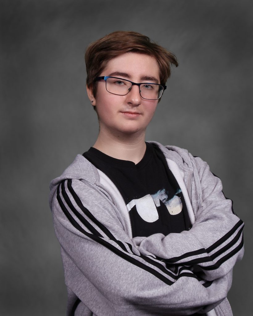A student with short brown hair and glasses stands confidently with arms crossed, wearing a gray hoodie and black T-shirt. Neutral gray background.