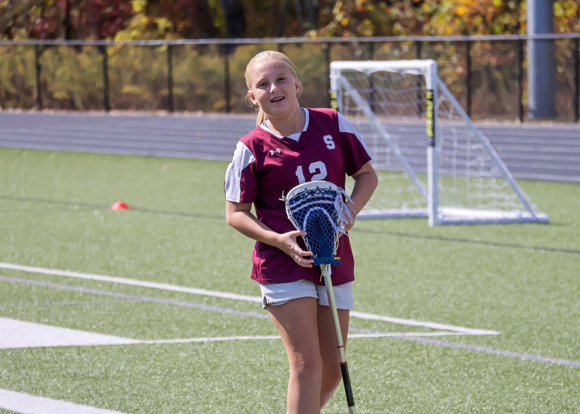 A middle school girl wearing a maroon lacrosse jersey and holding a lacrosse stick smiles while standing on a field with goalposts in the background.