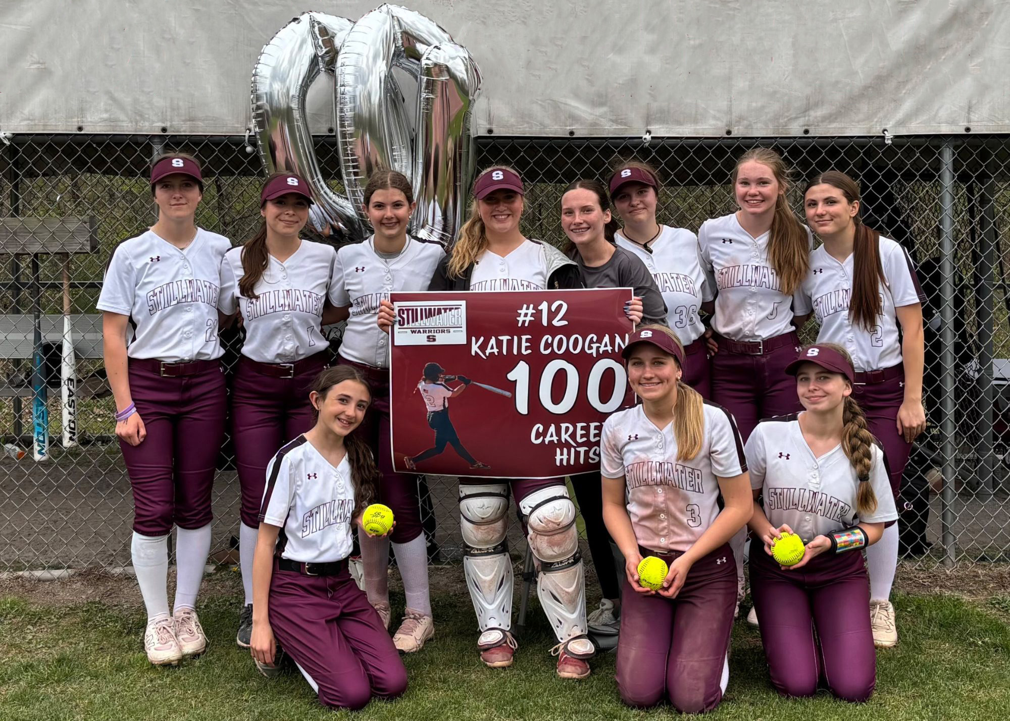 A group of softball players poses together, celebrating a milestone for player #12, Katie Coogan, holding a sign that reads "100 Career Hits." There are silver balloons in the background.