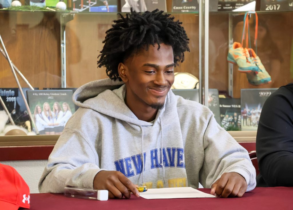 A young man with curly hair wearing a hoodie sits at a table, smiling and looking down at a sheet of paper.