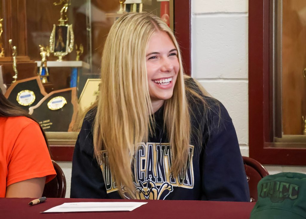 A teenage girl with long blonde hair is smiling, wearing a dark hoodie. She is seated at a table, and there are trophies displayed behind her.
