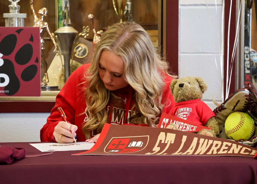 A young woman with long, wavy blonde hair is sitting at a table covered with a maroon cloth, writing on a piece of paper. She is wearing a red sweatshirt that reads "St. Lawrence." Next to her on the table are trophies and stuffed animals, including a bear and items related to softball.