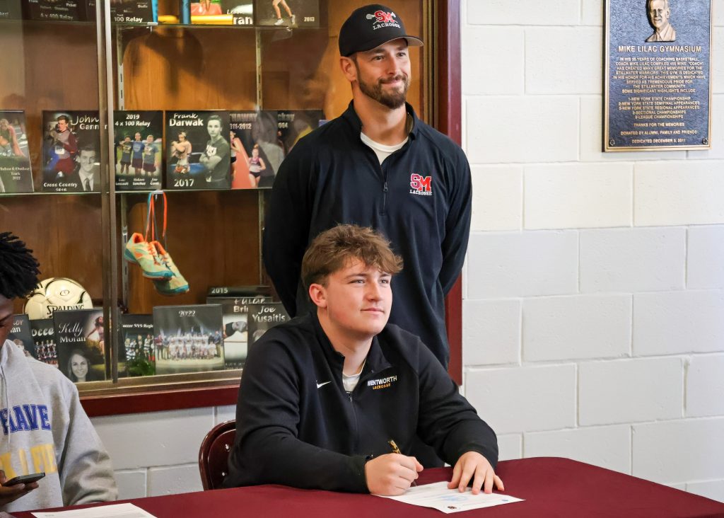 A student sitting at a table with a pen in hand, appearing to sign a document, while his coach stands behind him. The setting includes trophy cases in the background.