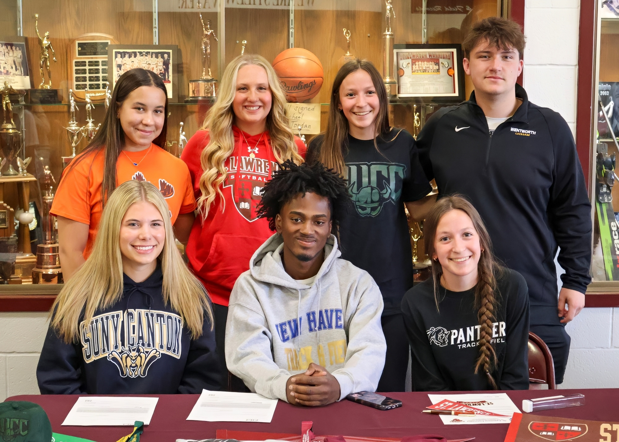 A group of seven students poses together, with two males and five females, in front of a display case filled with trophies. The male in the front center is sitting at a table with papers and a pen. The students are wearing various school apparel and smiling towards the camera.