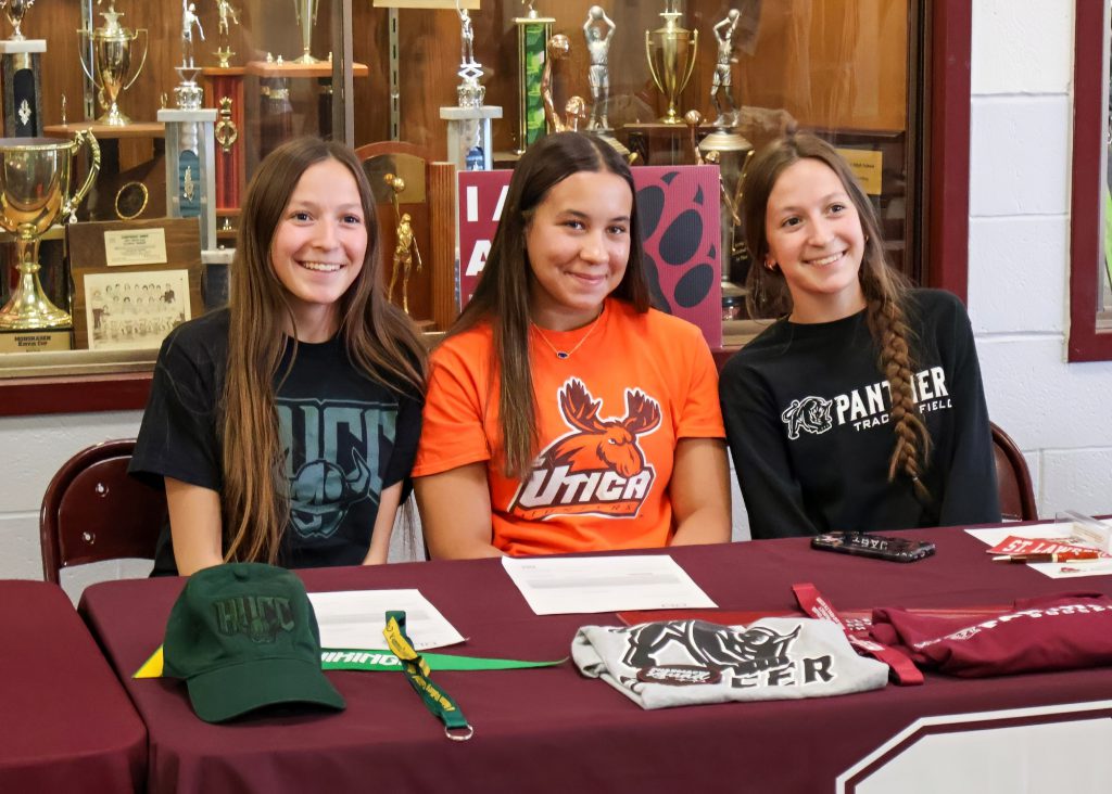 Three young women sitting at a table in front of a display of trophies, wearing shirts representing different universities. All are smiling towards the camera.