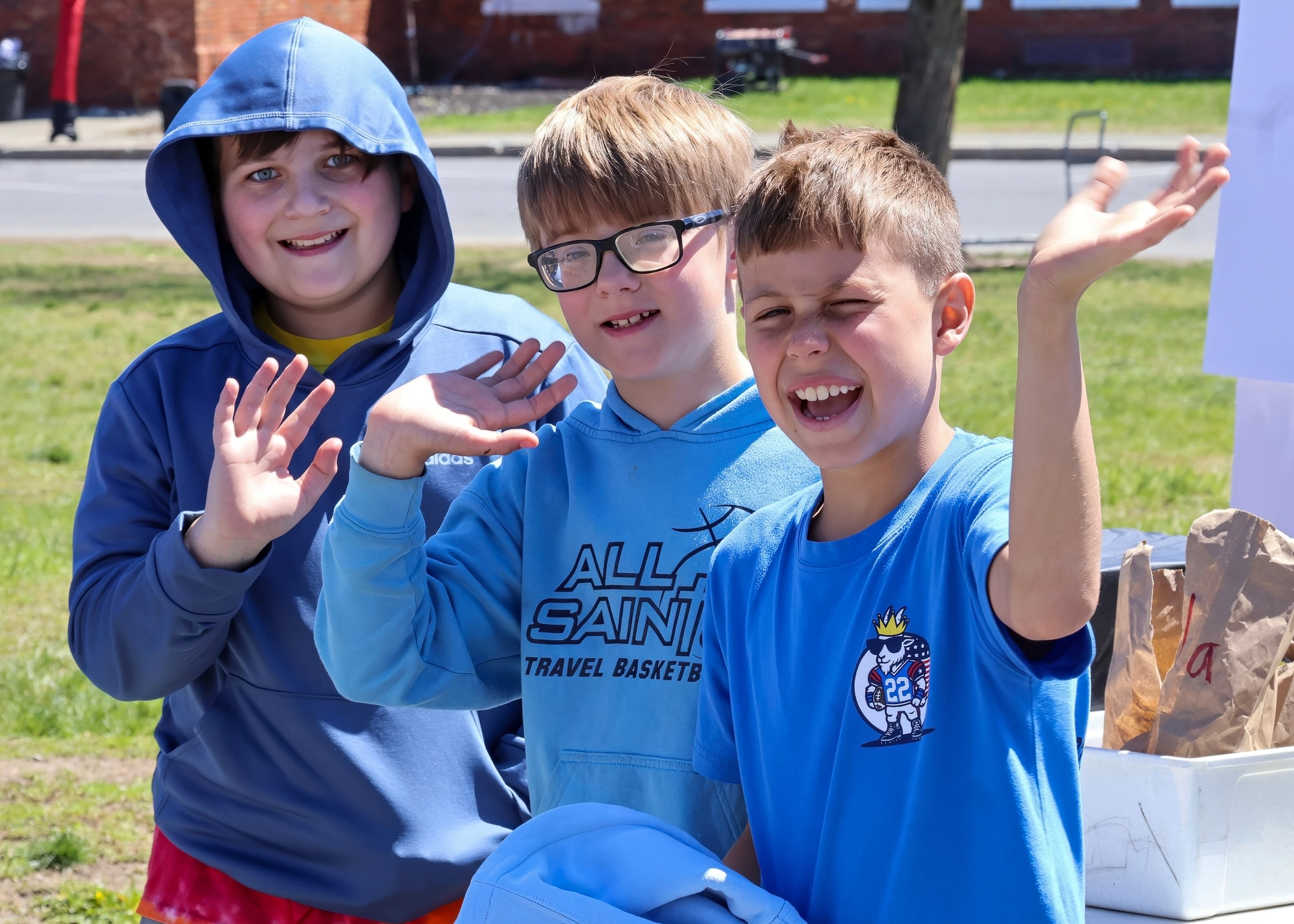 Three elementary-age boys smiling and waving at the camera while standing outdoors. The boy on the left is wearing a blue hoodie, the boy in the middle has glasses and is wearing a light blue sweatshirt, and the boy on the right is wearing a blue t-shirt.