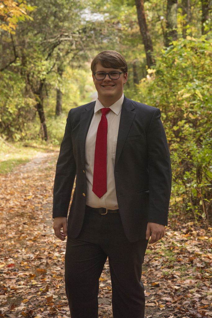 A young man in a dark suit and red tie stands smiling on a leaf-covered forest path. The setting is vibrant, with lush greenery and sunlight.