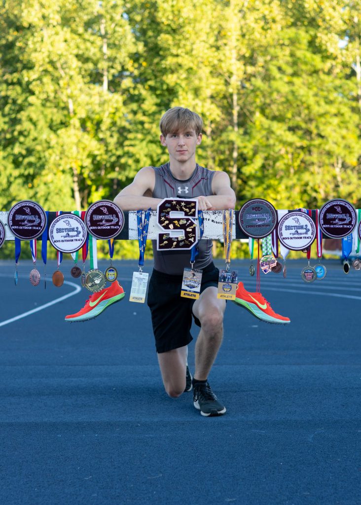 A young student athlete kneels on a track, surrounded by numerous medals and trophies displayed on a rack. Two orange running sneakers hang from the rack as well. Two backpacks sit on the ground, with trees in the background.