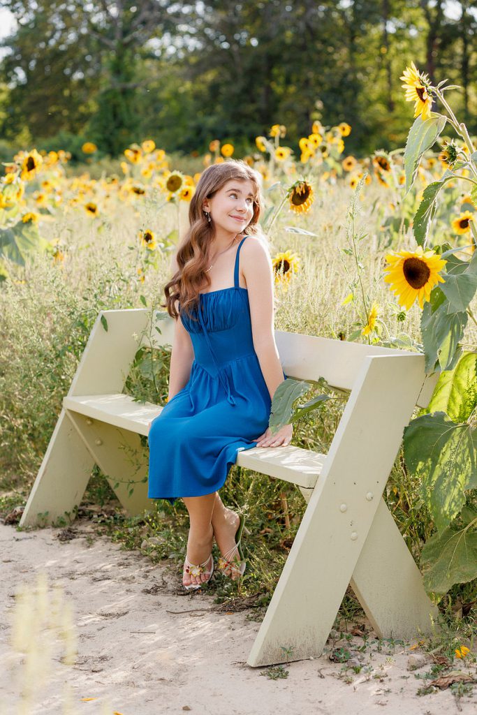 A young woman in a blue dress sits on a wooden bench in a vibrant sunflower field, looking to the side with a serene smile. Sunflowers and greenery surround her.