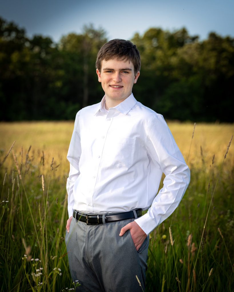 A young man stands in a sunlit field wearing a white collared button down shirt and gray pants, hands in pockets, smiling. Tall grass surrounds him against a forest backdrop.