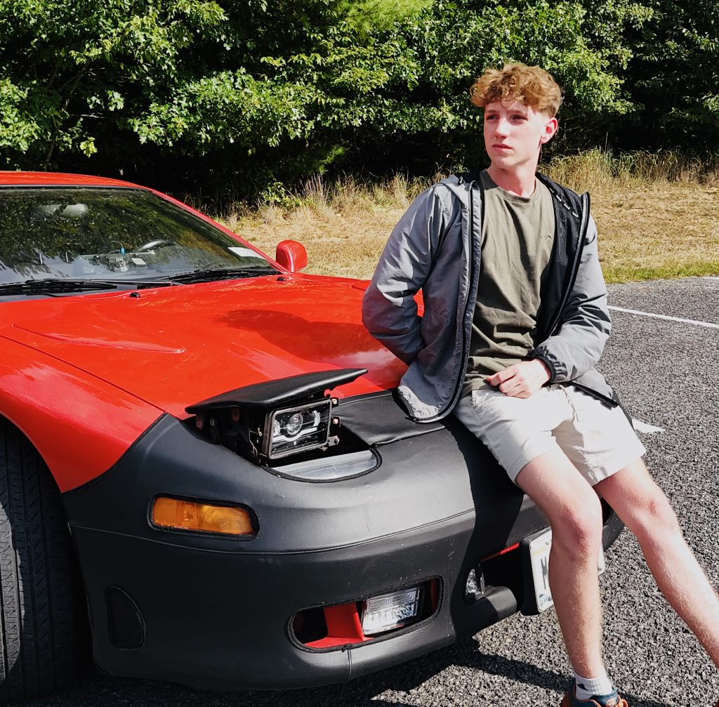 A young man leans on a vibrant red sports car, set against a background of lush greenery. He looks off to the left.