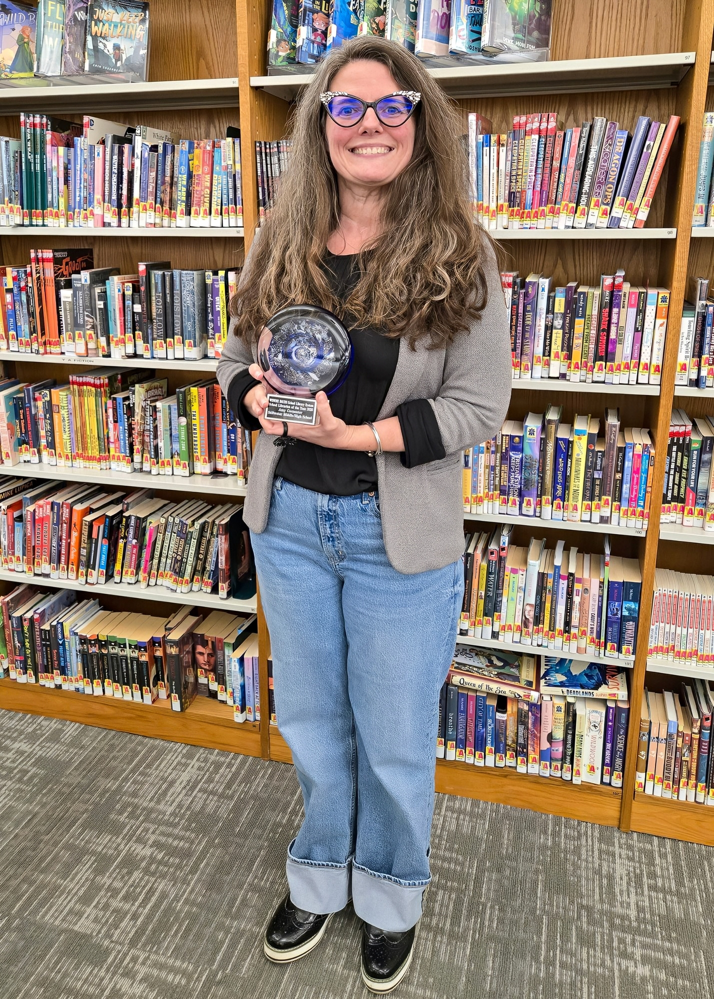 A woman with long, wavy hair wearing glasses and a gray cardigan stands in a library holding an award in front of a bookshelf filled with various books.
