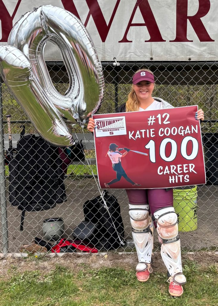 A young woman in a softball uniform holding a sign that celebrates her achievement of 100 career hits, with a large balloon number "100" in the background. The sign reads "#12 Katie Coogan 100 Career Hits."