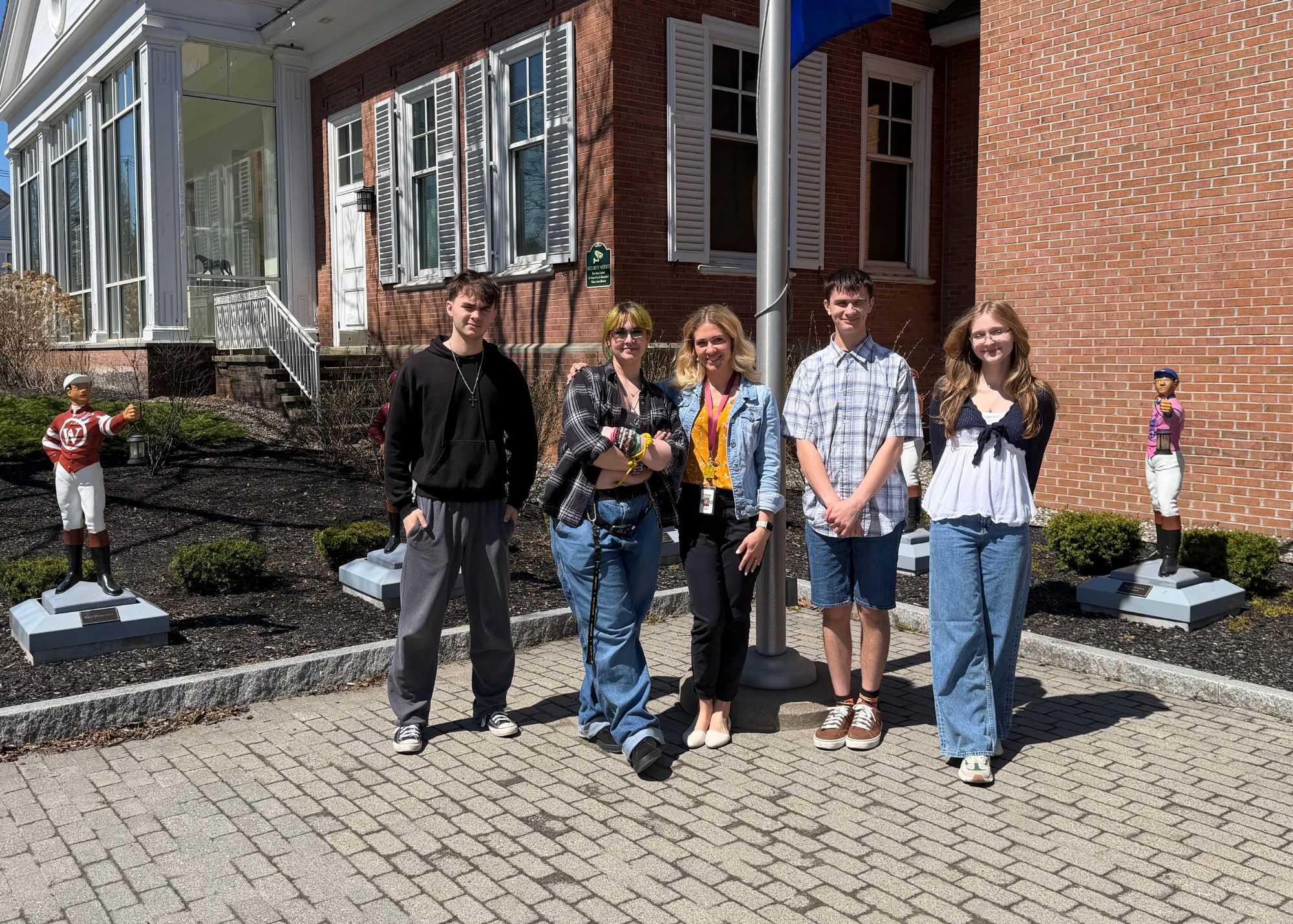 Five people stand in a row outside a brick building with white trim and windows, near small jockey statues on display in the background.