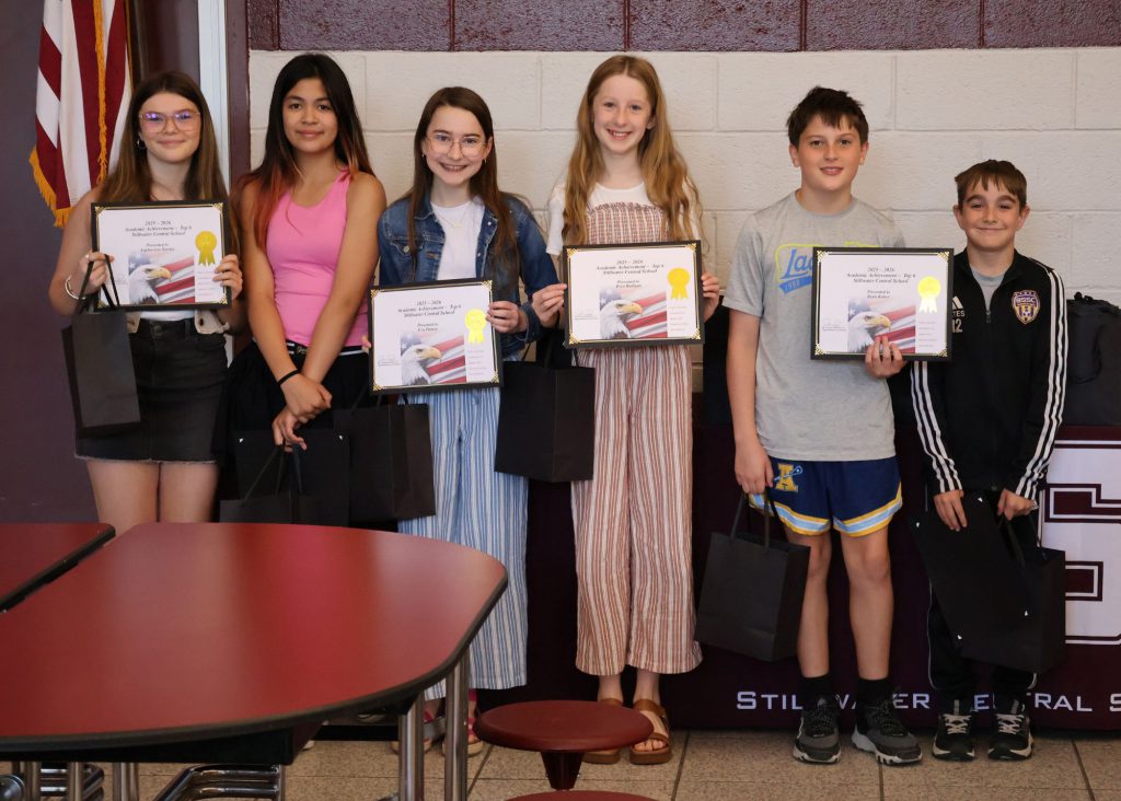 A group of six students holding certificates and gift bags, standing in front of a maroon/white wall with the Stillwater Central School District "S" logo in white. There are four girls and two boys.