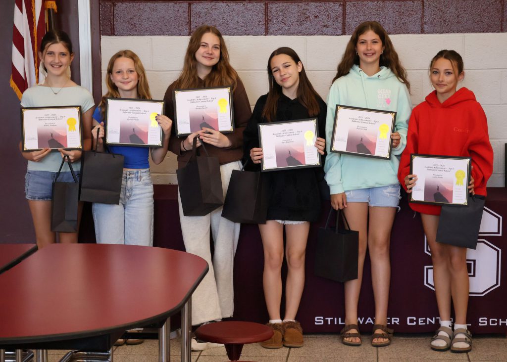 A group of six girls stands together. They are smiling and holding certificates and gift bags, standing in front of a maroon/white wall with the Stillwater Central School District "S" logo in white.