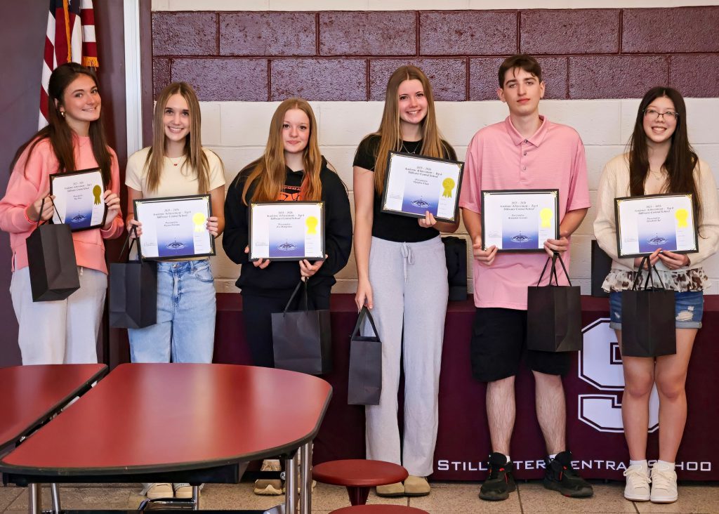A group of six students holding certificates and gift bags, standing in front of a maroon/white wall with the Stillwater Central School District "S" logo in white.