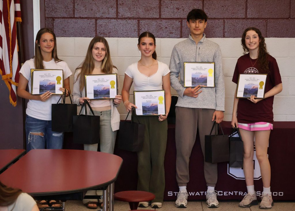 A group of five students holding certificates and gift bags, standing in front of a maroon/white wall with the Stillwater Central School District "S" logo in white. There are four girls and one boy.