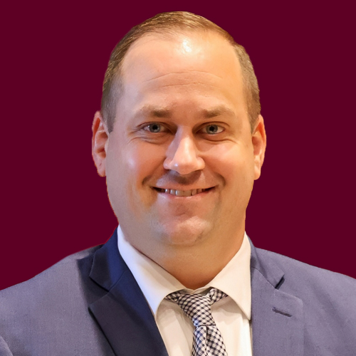 A man wearing a suit and tie, smiling at the camera. There's a maroon background.