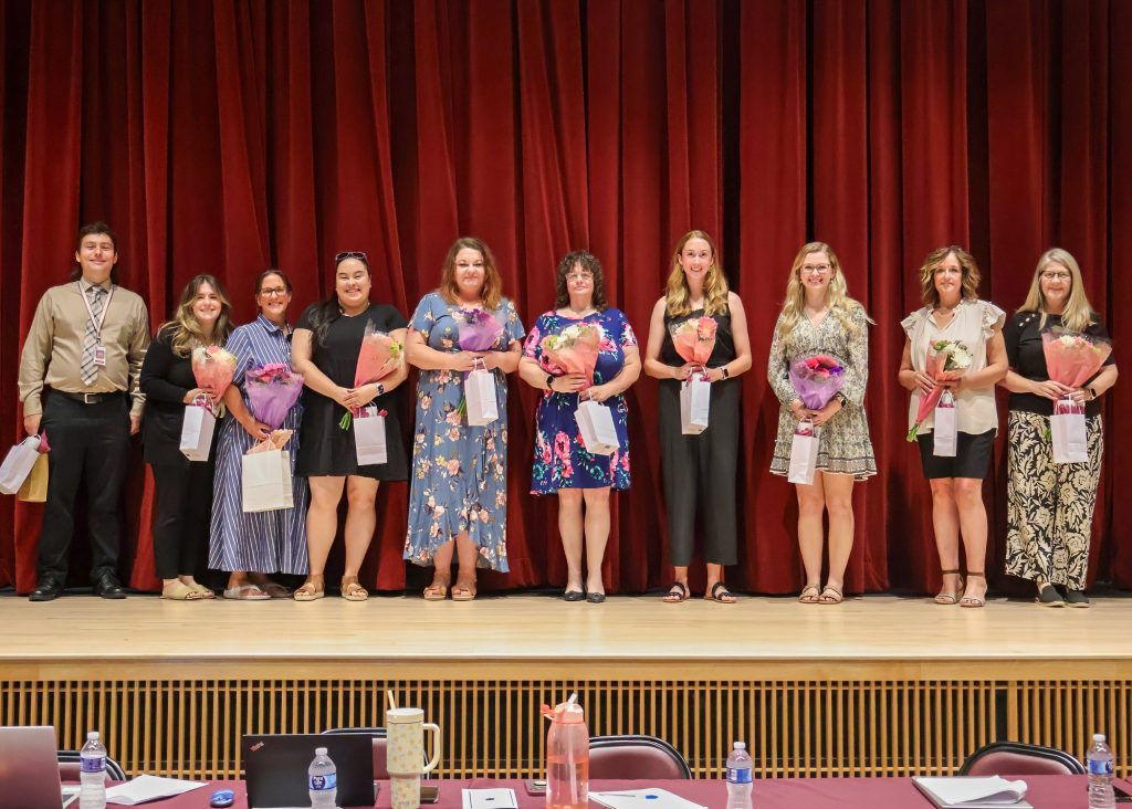 A group of ten individuals standing on a stage in front of a red curtain, each holding bouquets of flowers and white bags. They are all smiling for a group photo.