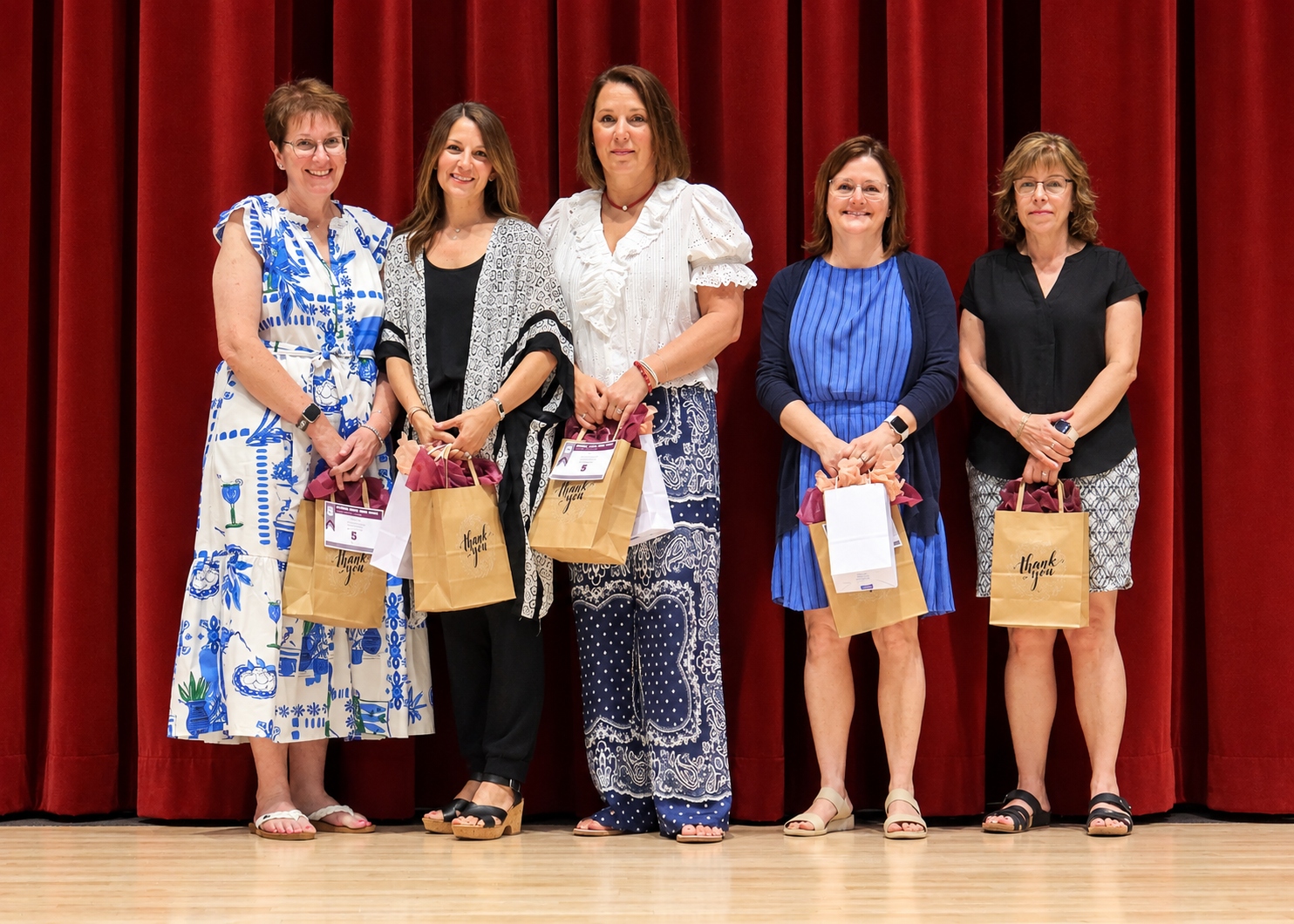 Five women standing together on a stage in front of a red curtain, each holding a small brown gift bag.