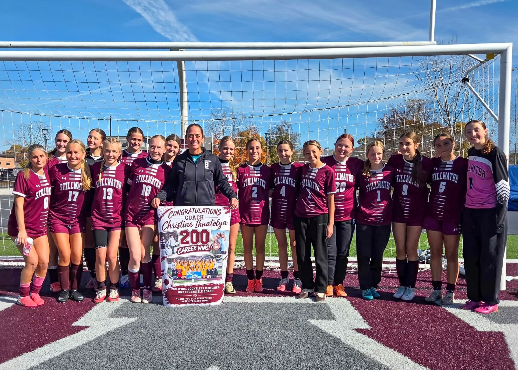 A girls’ soccer team poses in maroon jerseys beside a sign celebrating the coach's 200 career wins, smiling in front of a goal on a sunny day.