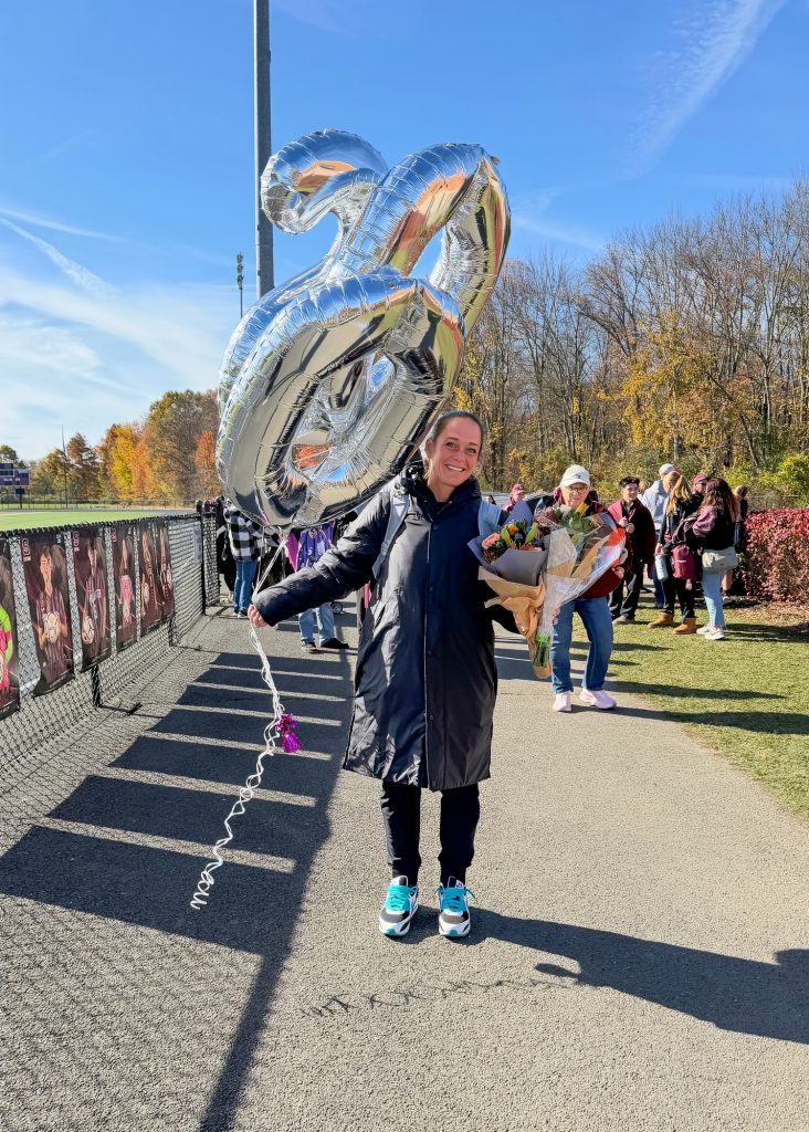 A smiling woman holds large silver number balloons outdoors on a sunny autumn day, with a gravel path, colorful trees and people gathered behind her.